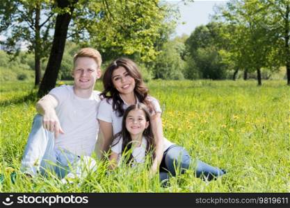 Happy smiling family of parents and daughter sitting on green grass in park. Family sitting in park