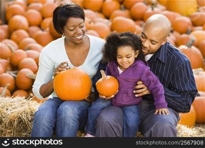 Happy smiling family sitting on hay bales and holding pumpkins at outdoor market.