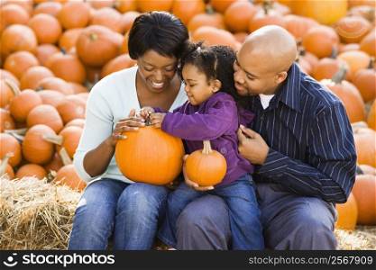 Happy smiling family sitting on hay bales and holding pumpkins at outdoor market.