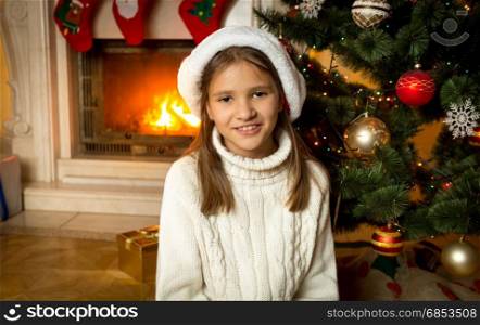Happy smiling girl in Santa hat sitting by the burning fireplace