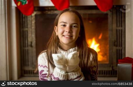 Happy smiling girl in sweater and gloves posing at burning fireplace and decorated Christmas tree