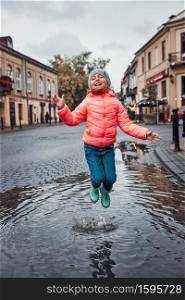 Happy smiling girl jumping in the puddle during walk in a downtown on rainy gloomy autumn day. Little girl jumping in the puddle on rainy gloomy autumn day
