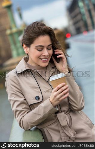 Happy, smiling, girl or young woman standing on Westminster Bridge, London, England, drinking coffee in a disposable cup and talking on a mobile cell phone