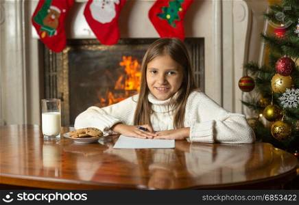 Happy smiling girl sitting by fireplace and writing letter to Santa Claus