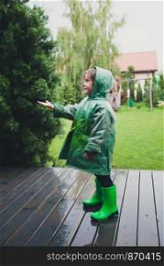 Happy smiling little girl standing on a porch catching raindrops wearing green raincoat
