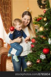 Happy smiling mother and baby boy decorating Christmas tree with baubles