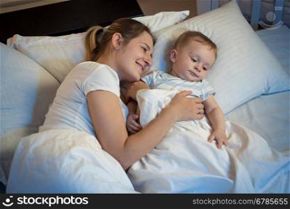 Happy smiling mother hugging her adorable baby in bed at night