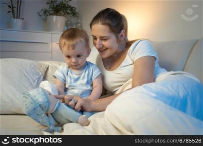 Happy smiling mother playing with her baby lying in bed at night and playing with toys