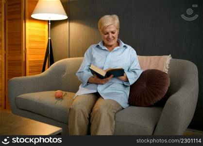 Happy smiling old woman reading book on sofa rest at home in evening. Leisure activity on retirement. Old woman reading book sitting on sofa