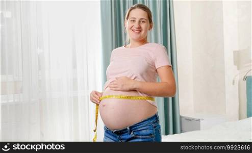 Happy smiling pregnant woman standing at window in bedroom and measuring big belly with measuring tape.. Happy smiling pregnant woman standing at window in bedroom and measuring big belly with measuring tape