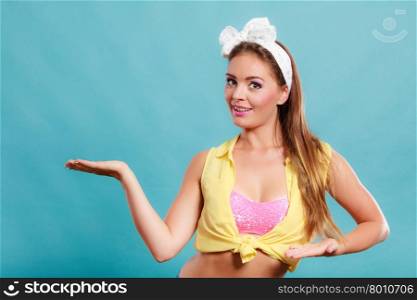 Happy smiling pretty pin up girl wearing hairband bow with empty hand palm for copy space. Attractive gorgeous young retro woman posing in studio on blue background.