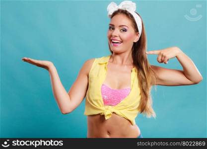Happy smiling pretty pin up girl wearing hairband bow with empty hand palm for copy space. Attractive gorgeous young retro woman posing in studio on blue background.