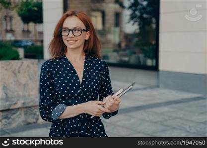 Happy smiling red-haired business woman holding laptop and notebook, looking aside while standing on city street outdoors. Female business professional with digital device near office building. Happy smiling red-haired business woman holding laptop , looking aside while standing on city street