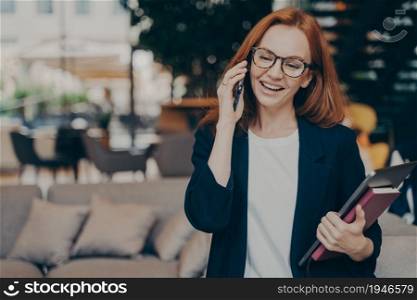 Happy smiling red haired business woman in optical eyewear enjoying telephone conversation, standing indoors with laptop in hand and talking on mobile phone with colleague or business partner. Happy smiling red haired business woman in optical eyewear enjoying telephone conversation