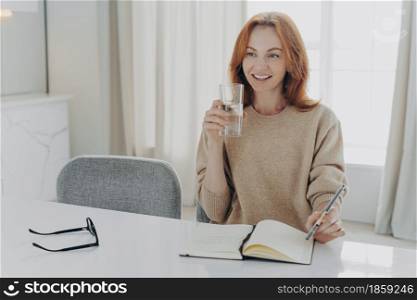 Happy smiling red haired woman housewife dressed casually with glass of pure mineral water enjoying morning at home, writing shopping list in notebook and planning her day, looking aside with smile. Happy red haired woman with glass of pure mineral water writing shopping list in notebook at home