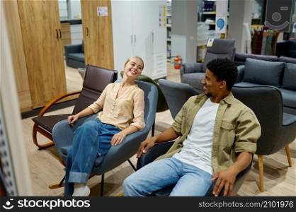 Happy smiling satisfied couple sitting on seat choosing new home chair in furniture store. Multiracial couple on chair in furniture store