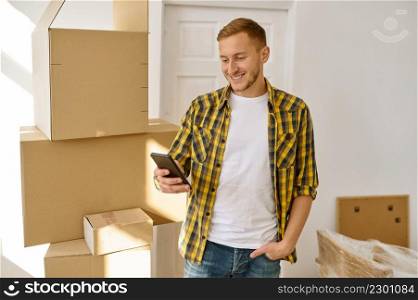 Happy smiling satisfied man having video call on mobile phone from new home stand among carton boxes. Man having video call from new home