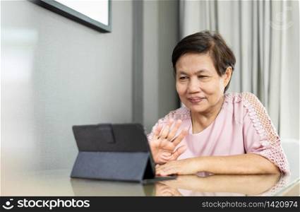 Happy Smiling Senior Asian woman making video call on digital tablet during quarantine and isolated at home. Elderly female in pink cloth waving hand while talking with people via internet technology.