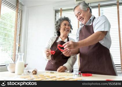 Happy Smiling Senior Lover man and woman baking in kitchen, Attractive Old couple enjoying holding red cookie cutter in heart shape while preparing and kneading dough at home together. Romance