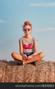 Happy smiling teenage girl sitting on a hay bale at sunset enjoying summer vacations in the countryside. Candid people, real moments, authentic situations