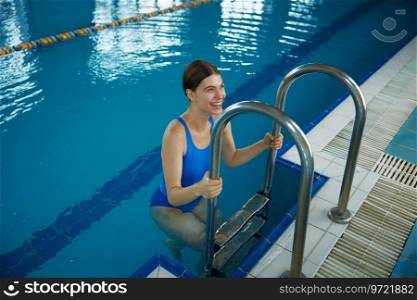 Happy smiling woman climbing out by steps of swimming pool. Training at poolside for healthy lifestyle concept. Happy smiling woman climbing out by steps of swimming pool