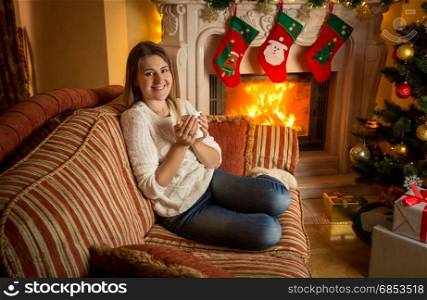 Happy smiling woman drinking tea on sofa at fireplace decorated for Christmas