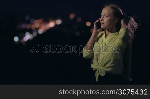 Happy smiling woman standing on viewing platform at night and talking on mobile phone. The wind is playing with her long beautiful hair. Blurred panorama of night city street on background