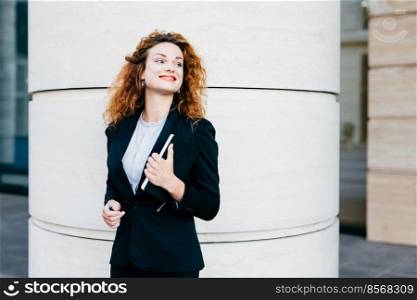 Happy smiling woman with red lips dressed formally, holding pocket book with pen looking aside. Glad businesswoman in white blouse, black jacket and skirt, looking elegantly. Female freelancer posing
