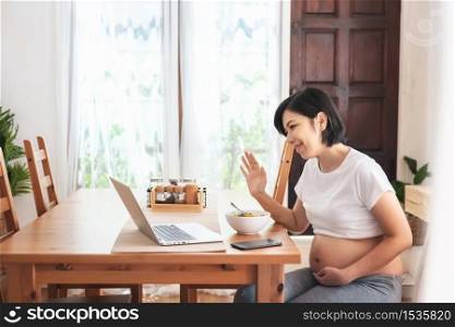 Happy smiling Young Asian Mother making video call online for talking and waving hand to family while her pregnancy at home. Joyful Beautiful Pregnant woman enjoy with laptop at table.