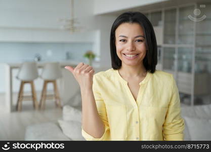 Happy smiling young female tenant pointing thumb finger aside on space for advertisement, standing at home. Online store sale, good deal offer. Real estate renting service advertising.. Happy female tenant pointing finger aside, advertising real estate renting service, standing at home