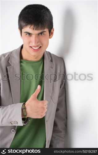 Happy smiling young man leaning against white wall and showing thumbs up
