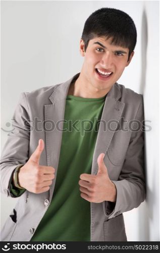 Happy smiling young man leaning against white wall and showing thumbs up