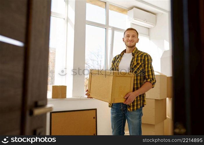 Happy smiling young man looking at camera carrying cardboard box during home or office relocation. Young man carrying cardboard box during relocation