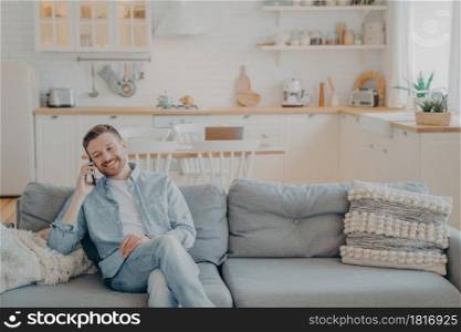 Happy smiling young man with stubble in casual clothes talking on phone with his friend while sitting with crossed legs on cozy couch, kitchen counter in blurred background. Communication concept. Young man talking on phone with his friend while sitting on couch