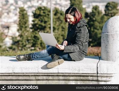 Happy smilling handsome redhead young woman sitting on a small wall and working on notebook - Concept Relaxing in nature.