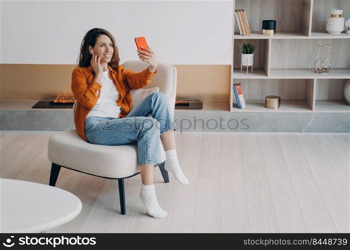 Happy spanish girl having video phone call at home and connecting earphones. Attractive young woman is using airpods. Technology using, leisure and communication concept. Beautiful modern interior.. Happy spanish girl having video phone call at home and connecting earphones.