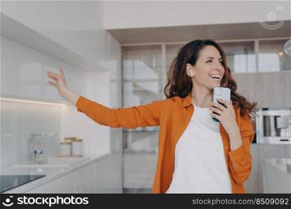 Happy spanish girl in airpods is singing with mobile telephone as with microphone. Woman having fun, relaxing and dancing. Application for music listening online. Housewife is singing song at home.. Happy spanish girl in airpods is singing with mobile telephone as with microphone.