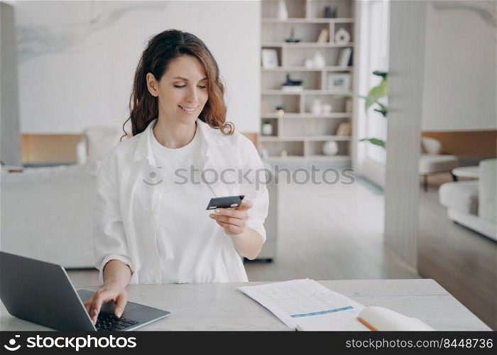 Happy spanish girl is holding credit card in front of pc. Young pretty woman is going to buy through internet. Girl is booking or purchasing online. E-commerce and financial transaction concept.. Happy spanish girl is holding credit card in front of pc. E-commerce and financial transaction.