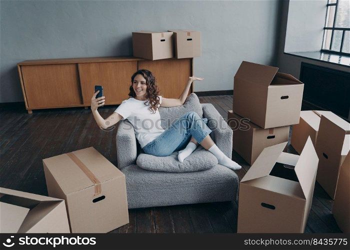 Happy spanish girl sitting in armchair and having video call on smartphone. Owner in new apartment. Successful young woman shows her dream house. Mortgage loan and independence concept.. Happy spanish girl sitting in armchair and having video call on smartphone. Owner in new apartment.