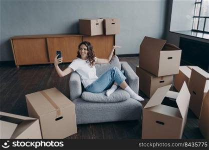 Happy spanish girl sitting in armchair and having video call on smartphone. Owner in new apartment. Successful young woman shows her dream house. Mortgage loan and independence concept.. Happy spanish girl sitting in armchair and having video call on smartphone. Owner in new apartment.