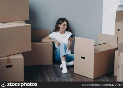 Happy spanish woman getting message from shipping service. Lady is packing boxes for the relocation. Girl is chatting on mobile phone and sitting on the floor. Easy moving concept.. Happy spanish woman getting message from shipping service. Lady is packing boxes for the relocation.