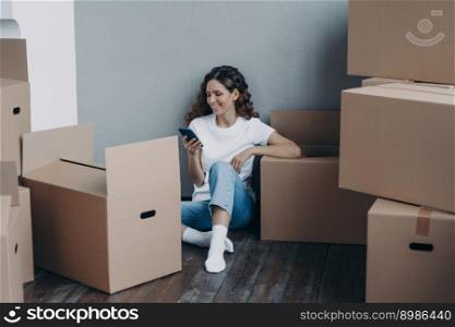 Happy spanish woman getting message from shipping service. Lady is packing boxes for the relocation. Girl is chatting on mobile phone and sitting on the floor. Easy moving concept.. Happy spanish woman getting message from shipping service. Lady is packing boxes for the relocation.