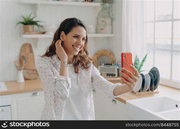 Happy spanish woman has video call on smartphone at kitchen. Girl is looking at camera of telephone and smiling. White scandinavian interior. Daylight through the window.. Happy woman has video call at kitchen. Girl is looking at camera of telephone and smiling.