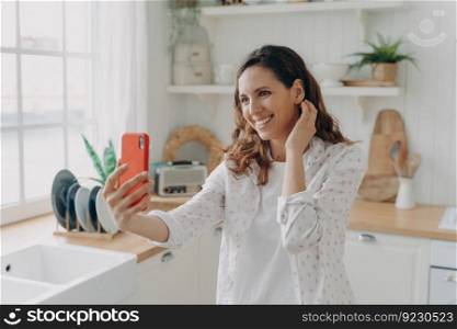 Happy spanish woman has video call on smartphone at kitchen. Girl is looking at camera of telephone and smiling. White scandinavian interior. Daylight through the window.. Happy woman has video call at kitchen. Girl is looking at camera of telephone and smiling.