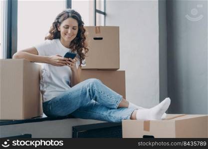 Happy spanish woman is sitting on windowsill of new home. Girl is unpacking boxes. Young lady is texting on phone and smiling. Joy of relocation concept.. Happy spanish woman is sitting on windowsill texting on phone and unpacking boxes.