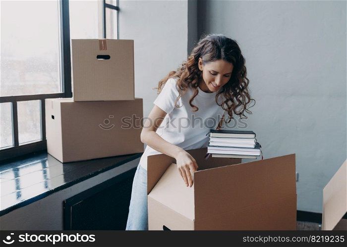 Happy spanish woman packing box with things, looking into cardboard package during moving, relocating in new home. Smiling girl putting her notebooks in carton. Relocation, real estate rental concept.. Happy woman packing carton box with things, relocating in new home. Moving, real estate rental