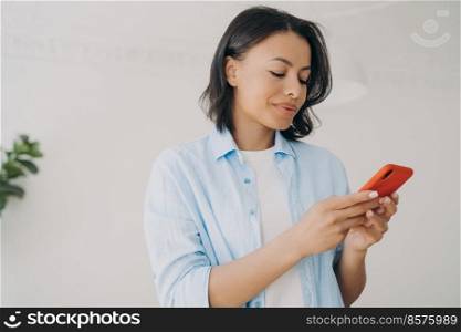 Happy spanish woman texting on smartphone and smiling. Young attractive lady indoor, copy space. Woman is chatting in social media on phone. SMS messages reading and typing. Communication technology.. Happy spanish woman is texting on smartphone and smiling. SMS messages reading and typing.