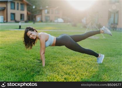 Happy sporty European woman stands in plank and raises legs, does stretching exercises, has morning workout alone, leads active lifestyle, dressed in sportswear, stays always fit and healthy