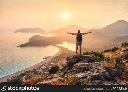 Happy sporty man with backpack and raised up arms standing and on the rock and looking at the seashore and mountains at sunset in summer. Man, sea, mountain ridges and orange sky. Oludeniz, Turkey. Young sporty man with backpack, seashore and mountains