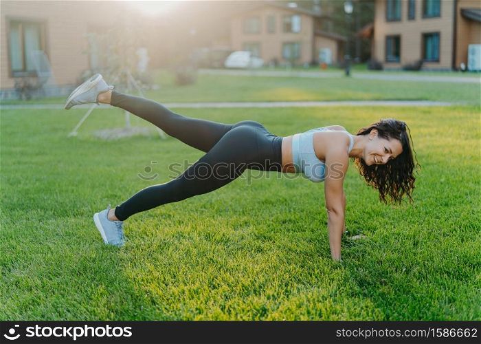 Happy sporty woman stands in plank pose, raises legs and does physical exercises to keep fit, dressed in top and leggings, goes in for sport regularly, enjoys nice summer day, poses on green grass